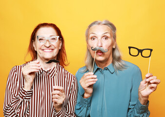 funny elderly female friends with fake mustache and glasses, laughs and prepares for party over yellow background