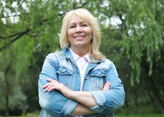 Obraz premium Stylish blonde woman in a denim jacket in a summer park. An elderly happy lady pensioner stands with her arms crossed.