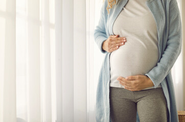 Close up of tummy of pregnant woman who gently supports him with her hands in anticipation of meeting baby. Cropped image of woman by window at home. Concept of motherhood, pregnancy and happiness.