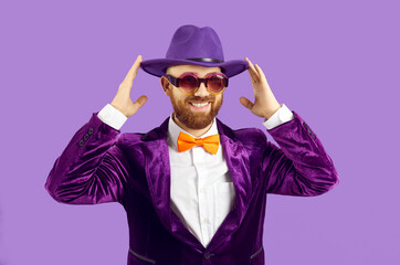 Funny smiling man in velvet jacket adjusts his hat while standing on purple studio background....