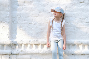 Fototapeta na wymiar Portrait of happy smiling child girl. Kid wearing white t-shirt and a summer hat. Light wall background. Street landscape.Copyspace