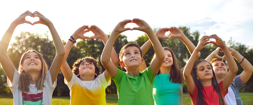 Group of happy good kind little children standing in row in green sunny summer park, looking at peaceful skies up above, raising hands, and doing heart gestures. Banner, header. Love and peace concept
