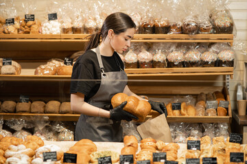 Young smiling girl with bread in the bakery, preparing pastry for sale in supermarket bakery...