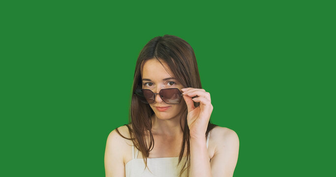young woman lowers her big sunglasses down her nose, looks up and puts her glasses back on chroma key. Green screen, portrait, close-up.