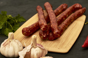 spicy smoked sausages on a cutting board next to red pepper garlic and herbs on a black background