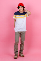 Portrait studio shot of Asian young handsome urban teenager fashion male model in street style outfit t shirt sport beige pants and red bucket hat standing posing look at camera on pink background