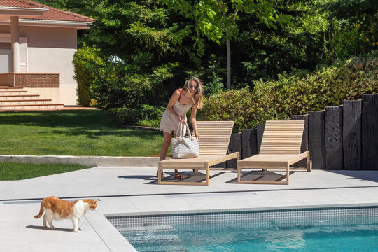 Young Blond Woman At The Pool In A Sun Lounge With Her White And Orange Cat Looking At The Water