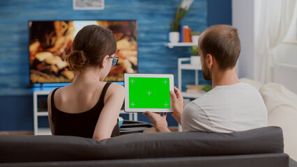 Man holding digital tablet with green screen watching online video content with girlfriend sitting on sofa. Couple looking at touchscreen device with chroma key enjoying influencer vlog.