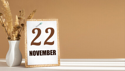 november 22. 22th day of month, calendar date.White vase with dead wood next to cork board with numbers. White-beige background with striped shadow. Concept of day of year, time planner, autumn month