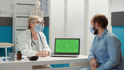 Medic and man with face mask looking at greenscreen on laptop, sitting at cabinet desk. People analyzing isolated copyspace background with blank mockup template and chromakey. Tripod shot.
