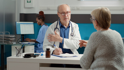 Male specialist writing health care report about senior patient at checkup visit apppointment. Taking notes about disease diagnosis to prepare recovery treatment on prescription paper.