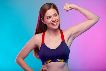 Fototapeta premium Happy young sporty woman in fitness sportswear flexes arm and demonstrates muscles, looking away. Female fitness portrait isolated on neon multicolor background.