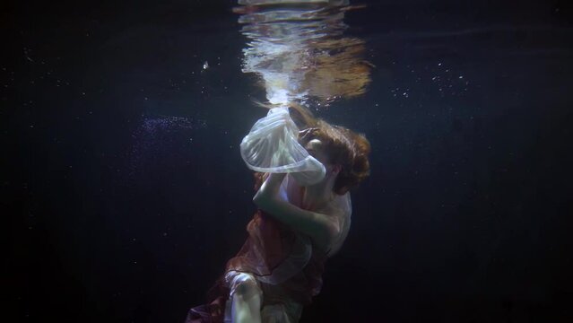 a red-haired woman hovers in the dark water. she is wrapped in white and red translucent fabrics