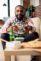 Smiling young person using tv remote contorl to switch channel and eating slice of pizza from fast food takeout delivery. Having fun with takeaway meal and beer while he watches movie.