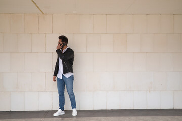 Handsome young man with beard, sunglasses, leather jacket, white shirt and jeans, next to a white wall, talking on his cell phone. Concept beauty, fashion, trend, smartphone, app, social networks.
