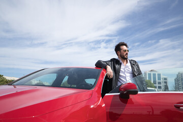 Handsome young man with beard, sunglasses, leather jacket and white shirt, leaning on the roof of his red sports car. Concept beauty, fashion, trend, luxury, motor, sports.