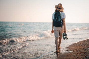 Young cheerful woman in a hat and sunglasses walking on the seashore with a backpack at sunset. Summer sea holidays
