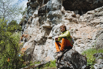 A portrait of a guy sitting on a stone in the mountains, a hike through the mountainous terrain, a tourist relaxing in the sun, a summer vacation in nature, a curly hipster blogger.