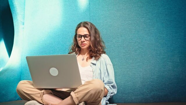 A young woman wearing glasses leading a video call with colleagues on her laptop while sitting cross-legged in a comfortable coworking space