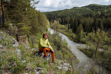 Hiking in the surrounding mountains, a person sits on a stone looking at the landscape, a tourist rests in the forest, hiking in the mountainous terrain, outdoor recreation. © Aleksey