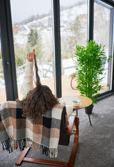 Curly young woman enjoying weekends inside contemporary barn house in the mountains. Female tourist sitting on chair in new cottage with panoramic windows.
