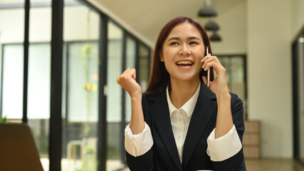 Happy asian female office worker talking on mobile phone, getting good news, celebrating hearing amazing news