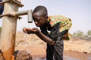Thirsty young black African boy leans forward to drink fresh and clean water with his bare hands...