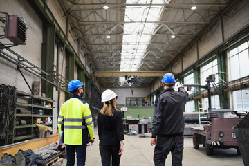 Rear view of manager supervisors and industrial worker in uniform walking in large metal factory hall and talking.