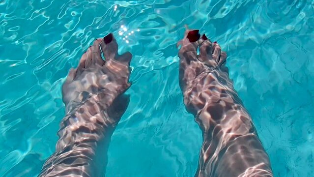 Clean blue pool water sparkles in the sun as it ripples and flows over a woman's feet with red nail polish casting shadows on her skin. High energy vacation image is refreshing and relaxing.