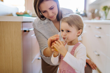 Young mother giving glass of water to drink to her little daughter in kitchen at home. - Powered by Adobe