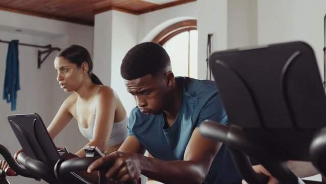 Focussed Black male and biracial female cycling on stationary bike. Having fun and determined at an indoor fitness gym.