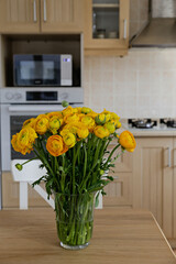 Bouquet of yellow ranunculus flowers in glass vase on the kitchen table. Stylish modern kitchen with small beige tile and wooden cupboard. Close up, copy space, background.