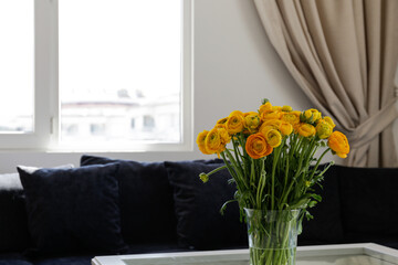 Close up shot of glass coffee table with bouquet of beautiful yellow ranunculus flowers in a vase and blue textile couch under the window on the background. Copy space, close up, natural light.