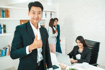 Portrait of a handsome CEO smiling, Portrait of a positive looking young business professional standing posing thumb up with coworkers talking in the background.