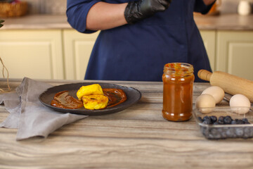 Female chef in a bright country kitchen.Dressed in a blue apron and black pants. Makes cheesecakes with salted caramel and fruit