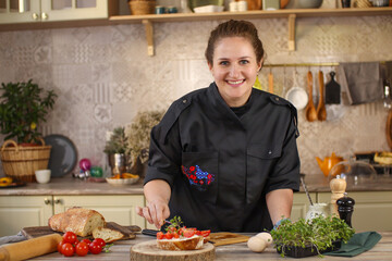 Female chef preparing breakfast in a bright country kitchen. He cuts the bread and spreads it with cheese. Kitchen table with flowers and kitchen board, bread, tomatoes, greens