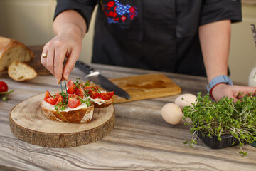 Female chef preparing breakfast in a bright country kitchen. He cuts the bread and spreads it with cheese. Kitchen table with flowers and kitchen board, bread, tomatoes, greens