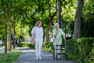 Caregiver with senior woman on walk with walker in park with shopping bag.