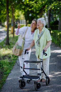 Caregiver with senior woman on walk with walker in park with shopping bag.
