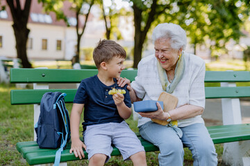 Great grandmother sitting on bench with her grandson and eating healthy fruit snack together,...