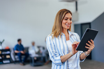 Fototapeta premium Side view of a businesswoman, holding and using a tablet while standing in the middle of the office.