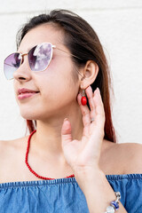 stylish young woman wearing jeans shirt, sunglasses and red jewelry at street, white wall background