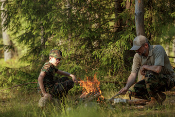 Father with son in military clothes is making campfire in the woods. The concept of adventure,...