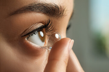 Young woman putting contact lens in her eye on blurred background, closeup - obrazy, fototapety, plakaty