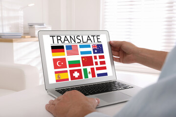 Translator using modern laptop with images of different flags on screen at white table indoors,...