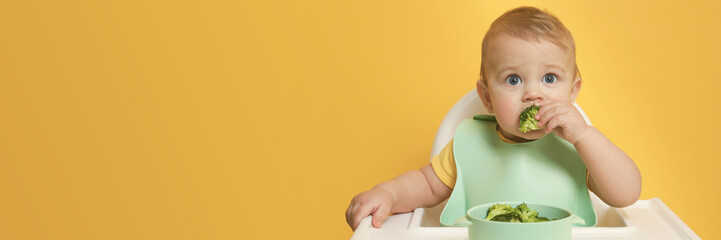 Cute little baby wearing bib while eating on yellow background, space for text. Banner design - obrazy, fototapety, plakaty