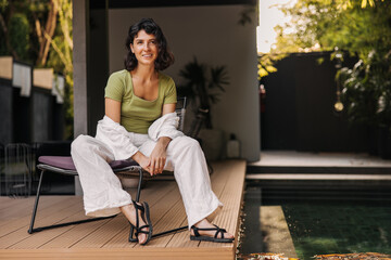 Obraz premium Attractive young caucasian brunette woman looks away, rests sitting on chair in courtyard of house. Girl wears T-shirt, shirt, pants and sandals. Summer vacation concept