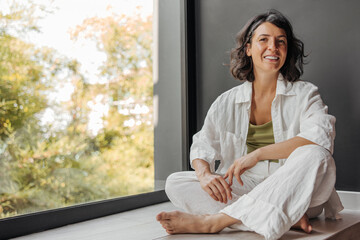 Cute young caucasian girl looks at camera sitting on floor near window in sunny weather. Brunette wears white shirt and pants. Lifestyle, female beauty concept