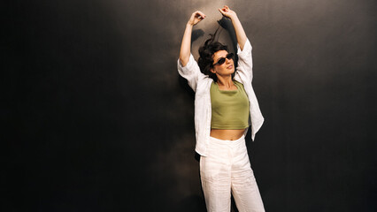 Relaxed caucasian young woman stretching arms above her head standing on black background with space for text. Brunette wears sunglasses and casual clothes. Rest time concept