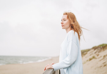 Fototapeta na wymiar Portrait of young red haired woman in light blue sweater on sand beach by sea in storm
